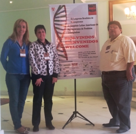 três pesquisadores posam para foto com banner do congresso ao fundo